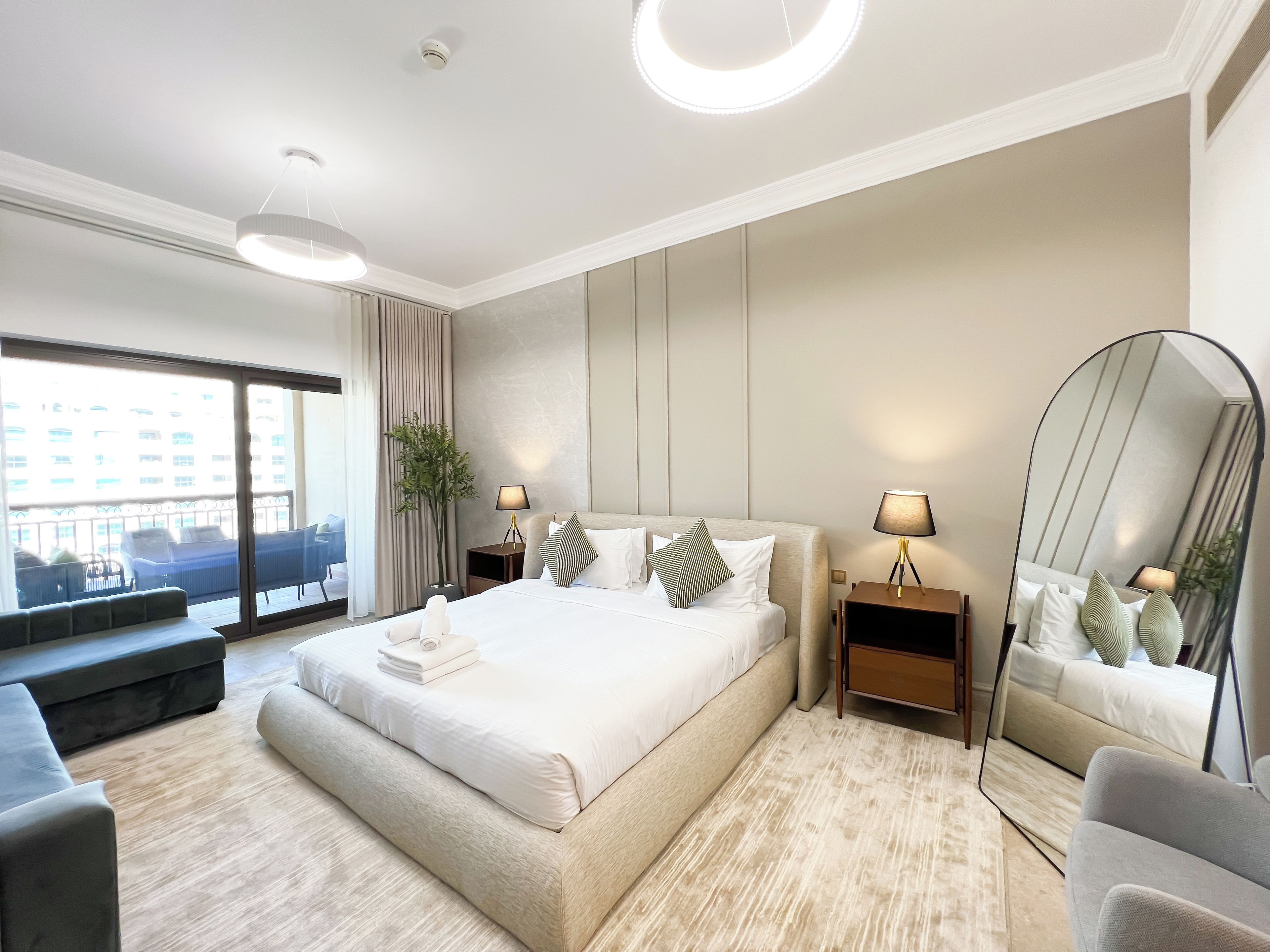 A modern hotel room with a large bed, a sitting area with a bench, a large mirror, and a view of the city through the window.