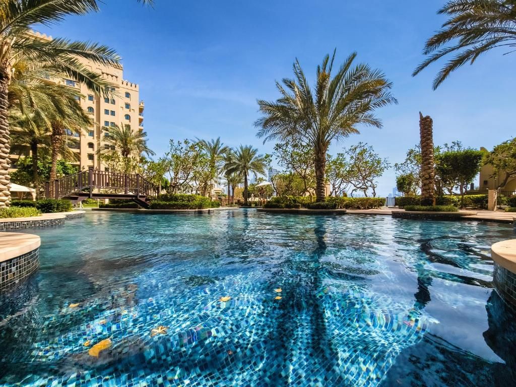 A swimming pool with clear blue water, surrounded by palm trees and a building with multiple floors.