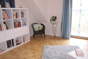 Een kamer met een boekenkast, een stoel, een tafeltje met een plant en een raam met gordijnen.