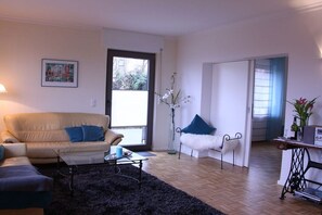 Een woonkamer met een bank, fauteuil en salontafel. Er is een deur naar een andere kamer, een raam met uitzicht, en een vaas met bloemen.