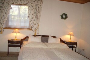 Une chambre à coucher avec un lit double, deux tables de chevet avec des lampes, une fenêtre ornée de rideaux à motifs floraux et une couronne décorative accrochée au mur.