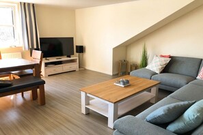 A modern living room with a flat-screen TV, a wooden coffee table, a grey sofa, and a dining area with a bench and table. - Holiday apartment in Binz with swimming pool (Binz)