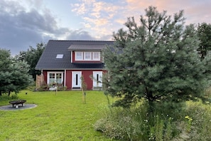 A two-story house with a red facade and a dark roof, surrounded by a well-maintained lawn and a large evergreen tree. - Holiday apartment with 1 bedroom (Pruchten)
