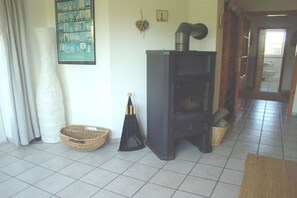 A room with a tiled floor, a black wood-burning stove, a framed picture on the wall, and a door leading to another room. - Spacious holiday apartment in Petersdorf (Petersdorf\/Fehmarn)