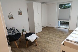 Ein Schlafzimmer mit einem Bett, einem Kleiderschrank, einem Sessel, einem kleinen Tisch mit einer Vase und einem Fenster mit Jalousien.