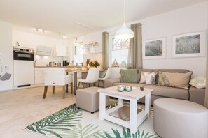 A modern living room with a sofa, ottoman, and coffee table, a kitchen area with white cabinets and appliances, and artwork on the walls. - Holiday apartment in Zingst with Wi-Fi (Zingst)