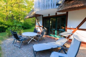 Eine Terrasse im Freien mit einem runden Tisch und Stühlen, einem Liegestuhl und einer überdachten Laube mit einem Reetdach.