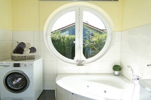 A bathroom with a washing machine, a bathtub, and a window with a view of a house and trees. - Luxury holiday home lighthouse (Bohnert)