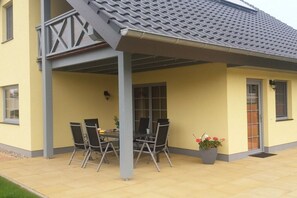 A covered outdoor seating area with a table and chairs, a potted plant, and a building with a tiled roof. - Holiday home with 4 bedrooms (Sternberg)