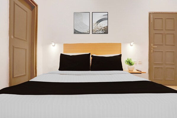 A bed with a wooden headboard, two black pillows, a white bedspread, and a black throw blanket. There are two wall-mounted light fixtures on either side of the bed, and a potted plant on a bedside table.