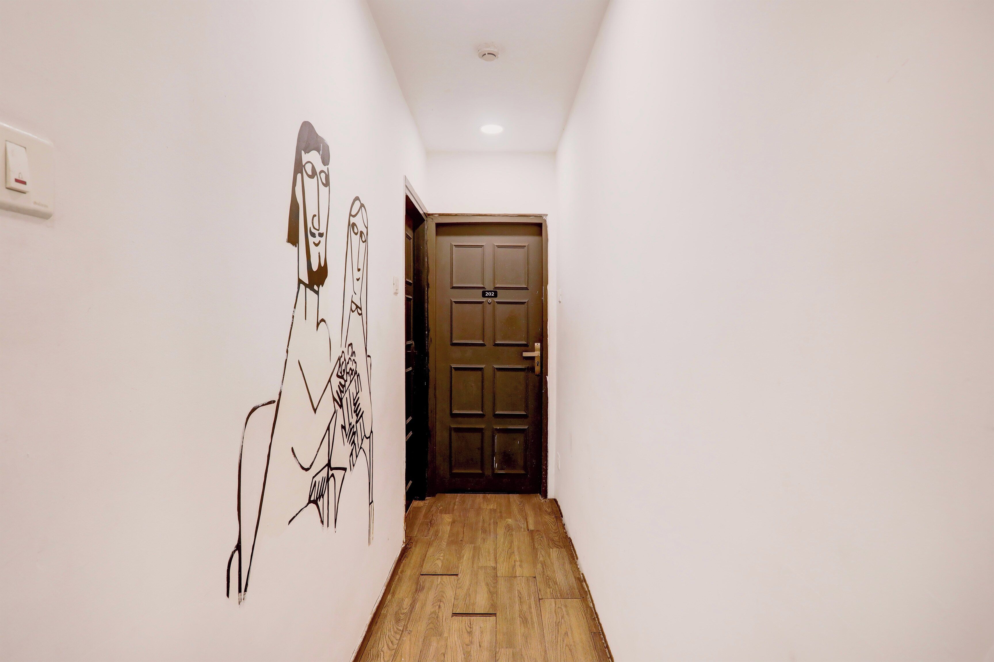 Un pasillo estrecho con suelo de madera, una puerta marrón con el número 202, y una pared con un gran dibujo abstracto.