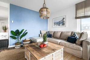 A living room with a blue accent wall, a wooden coffee table with fruit, a beige sofa, and a hanging wicker light fixture. - Holiday apartment at the marina (Damp)