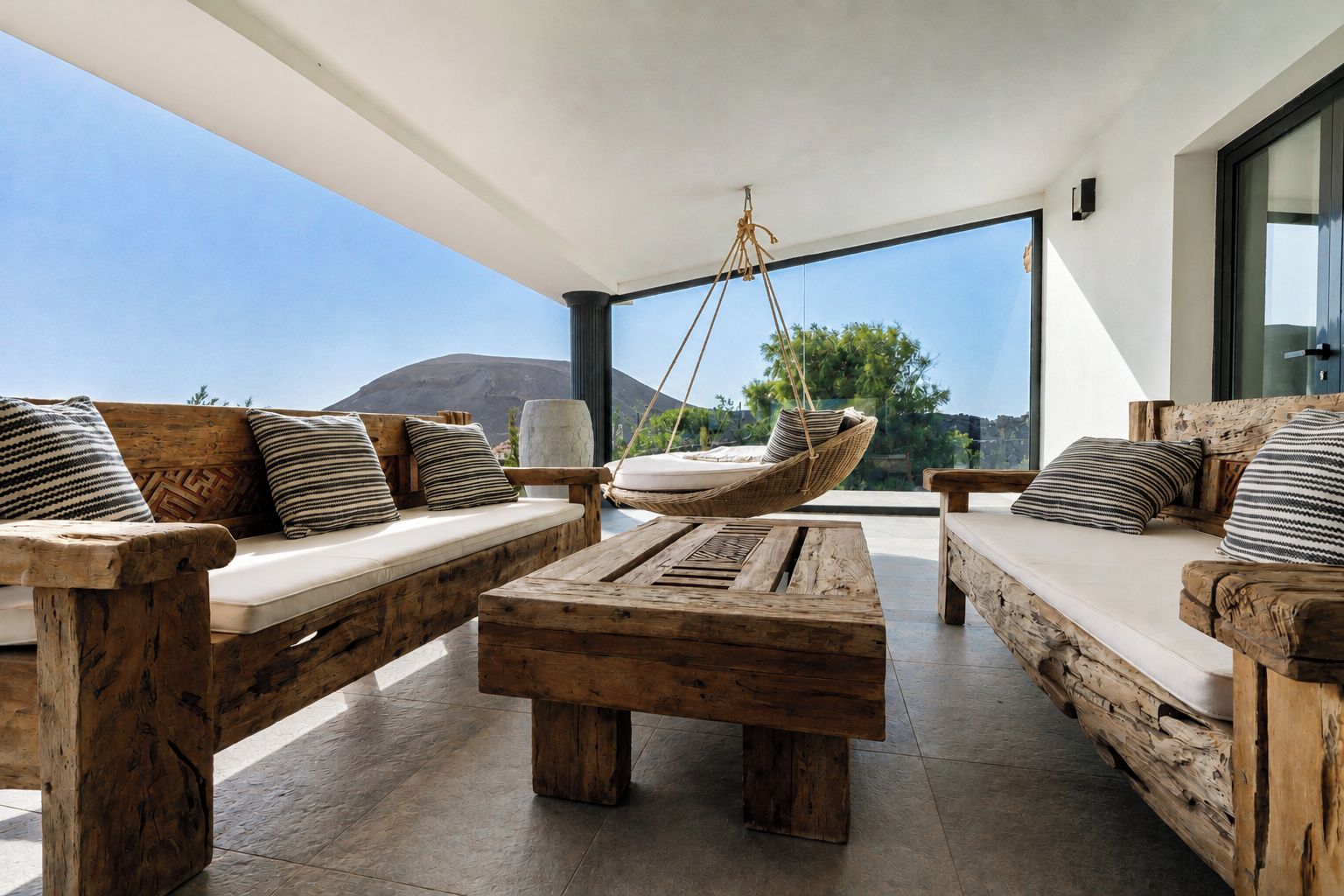 Un espacio exterior moderno con mobiliario de madera y una silla colgante, con vistas a un paisaje montañoso.