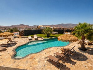 A swimming pool with lounge chairs and a thatched umbrella, surrounded by a stone patio and a view of mountains.