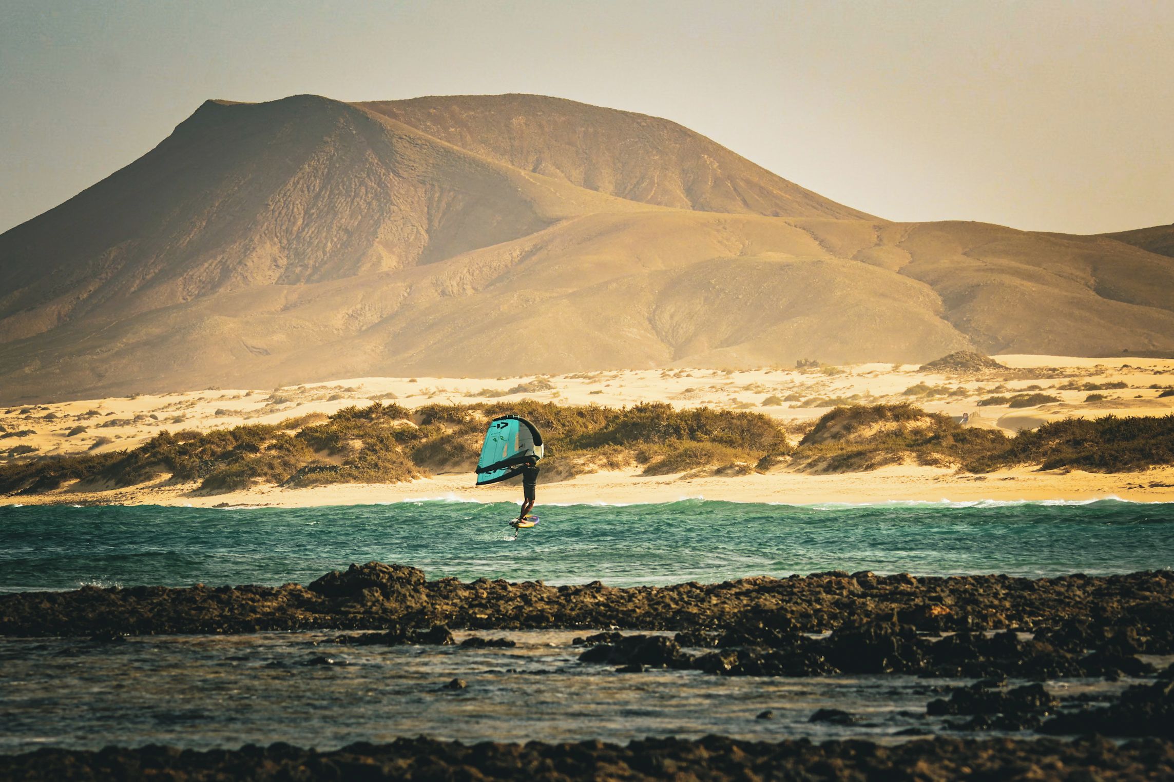 Una persona practica windsurf en una playa con una montaña al fondo.