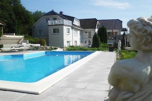 En swimmingpool med en klar blå vandoverflade, omgivet af en belagt gangsti. I baggrunden ses to huse med synlige altaner og en statue af en figur.