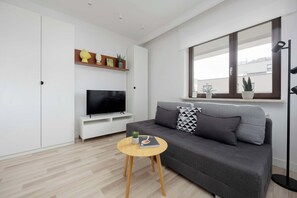 Ein modernes Wohnzimmer mit grauem Sofa, einem Flachbildfernseher, einem kleinen Holztisch und einem Fenster mit Blick auf Gebäude.