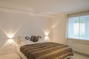 Quarto com cama, duas luminárias de cabeceira, janela com persianas e radiador.