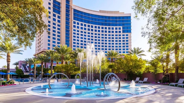 Un hotel con un amplio área de piscina, con múltiples chorros de agua y fuentes, rodeado de palmeras y sillones de descanso.