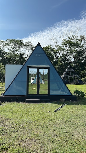 A modern glass pyramid structure with a reflective surface, surrounded by a grassy area and trees. - Pachamama Pyramid 5 (La cruz)