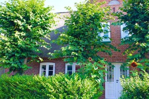 Un edificio de ladrillo con una puerta y ventanas blancas, rodeado de abundante follaje verde.