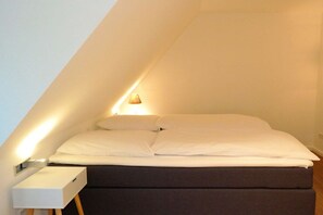 A single bed in a room with a sloped ceiling, a bedside table, and a wall-mounted lamp. - 4 Caymans at Boldix House (Wyk-Boldixum)