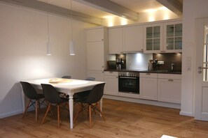 A modern kitchen with white cabinetry, a central island, and a dining area with a white table and black chairs. - 4 Caymans at Boldix House (Wyk-Boldixum)