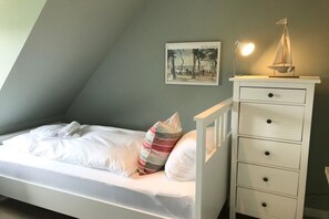 A single bed with white bedding and striped pillows, a white dresser with drawers, a framed picture on the wall, and a small sailboat model on top of the dresser. - View of the dike in Nieblum (Nieblum)