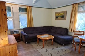 A living room with a dark-colored sofa, a wooden coffee table, and a dining table with chairs. There is a painting on the wall and a window with curtains. - Otto House - Holiday Apartment 1 (Wyk auf Föhr)