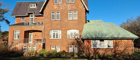A two-story brick house with a green roof, a balcony on the upper floor, and a well-maintained lawn.