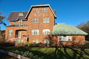 A two-story brick house with a green roof, a balcony on the upper floor, and a well-maintained lawn. - Otto House - Holiday Apartment 1 (Wyk auf Föhr)