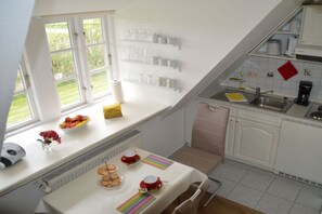 Een keuken met een raam, witte kastjes, een aanrecht en een tafel gedekt met een fruitschaal, waterkoker en kopjes.