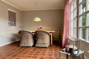 A room with a dining table, two wicker chairs, a hanging light fixture, and a window with curtains. - Holiday home in Nieblum with swimming pool (Nieblum)