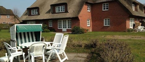 Een traditioneel huis met rieten dak en bakstenen muren, een terras met witte kunststof meubels, en een groen zonnescherm.