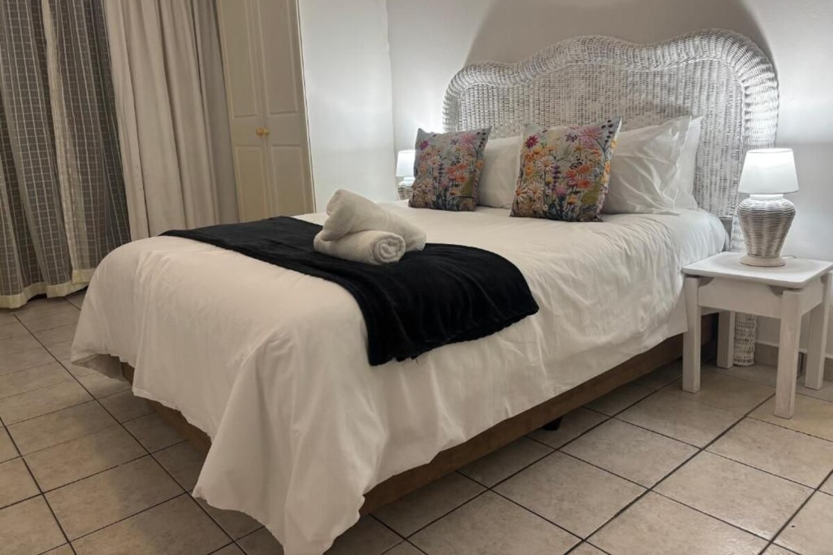 Ein ordentlich bezogenes Bett mit weißer Daunendecke, einer schwarzen Tagesdecke und blumigen Kissen. Neben dem Bett steht ein weißer Nachttisch mit einer Lampe und einem Korbwägelchen.