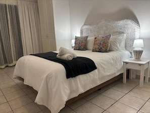 Ein ordentlich bezogenes Bett mit weißer Daunendecke, einer schwarzen Tagesdecke und blumigen Kissen. Neben dem Bett steht ein weißer Nachttisch mit einer Lampe und einem Korbwägelchen.