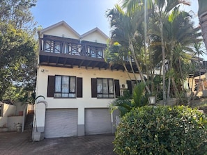 Ein zweistöckiges Gebäude mit Balkon, Garage und einem gepflegten Garten mit Palmen.
