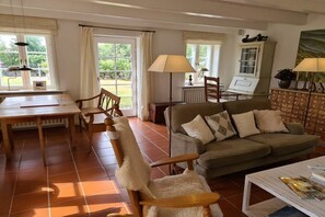 Ett vardagsrum med ett matbord i trä, en soffa och ett soffbord. Det finns två lampor, ett skåp och ett fönster med gardiner.