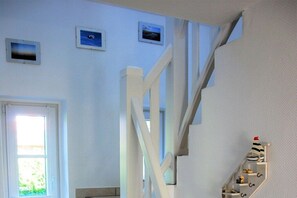 A staircase with white railings leading to a landing, a window with a view of greenery, and a shelf with various items. - Holiday home in Nieblum with swimming pool (Nieblum)