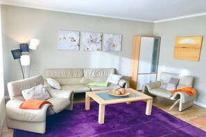 Ein Wohnzimmer mit einem beigefarbenen Sofagarnitur, einem Couchtisch und einem violetten Teppich. An den Wänden hängen drei Kunstwerke, und eine Stehlampe steht im Raum.
