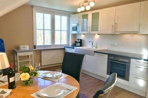 A modern kitchen with white cabinets, a microwave, a coffee maker, and a dining area with a table set for two. - Haus Heike - Holiday Apartment 1st Floor (Utersum)