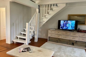 Un salon avec un escalier blanc, un téléviseur posé sur un meuble en bois et un magazine sur une table basse.