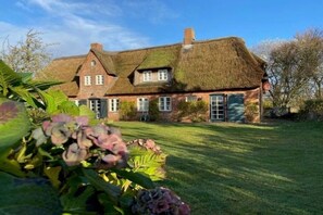 Une maison traditionnelle en briques, avec un toit de chaume, entourée d’une pelouse bien entretenue et de plantes en fleurs.