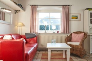 A cozy living room with a red leather sofa, a white coffee table, a wicker chair, and a window with curtains. - Holiday home with 1 bedroom (Wrixum auf Föhr)