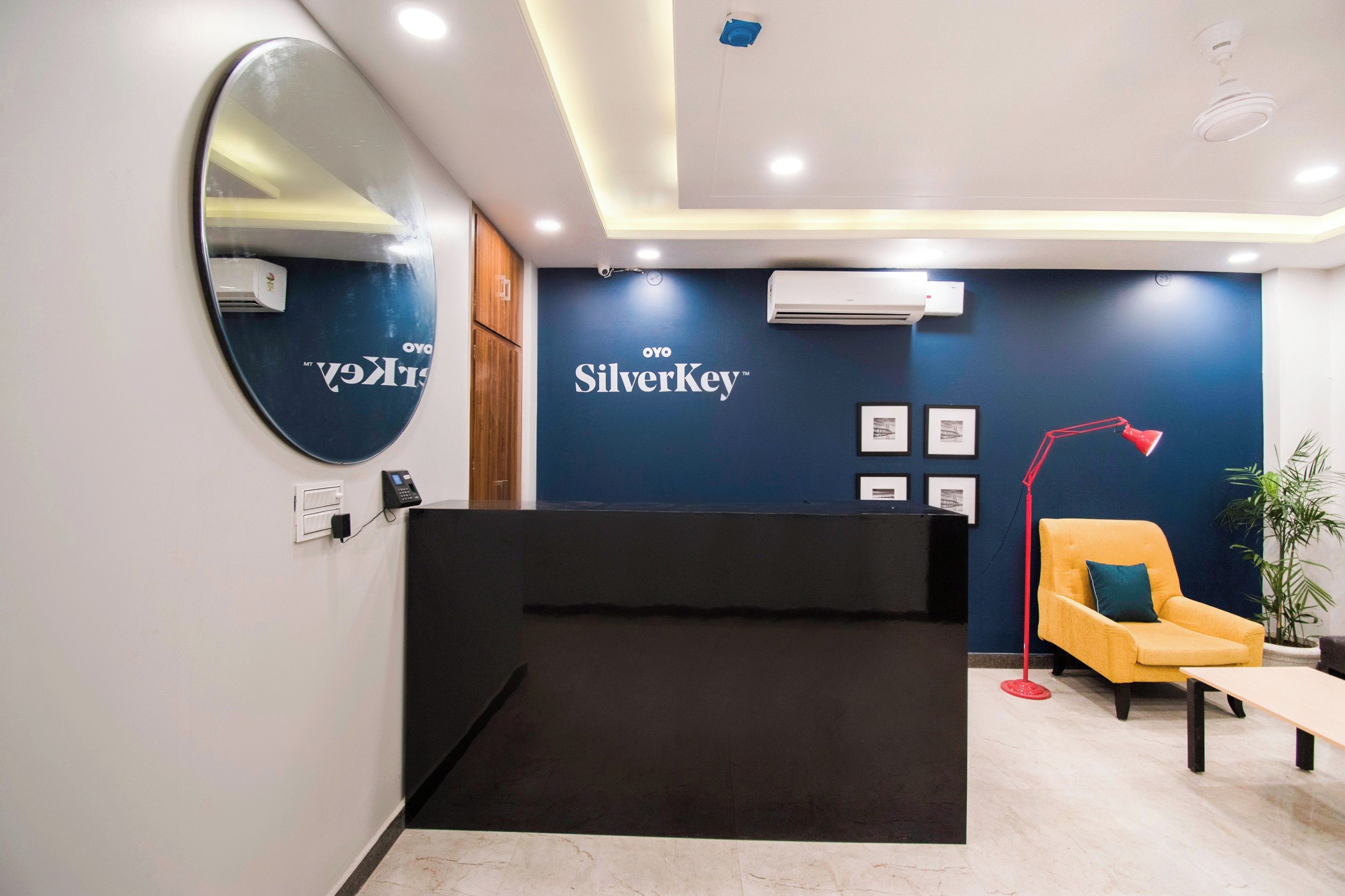 Un área de recepción con un mostrador negro, una pared azul con el logotipo 'Oyo SilverKey', un sillón amarillo y una lámpara de pie roja.