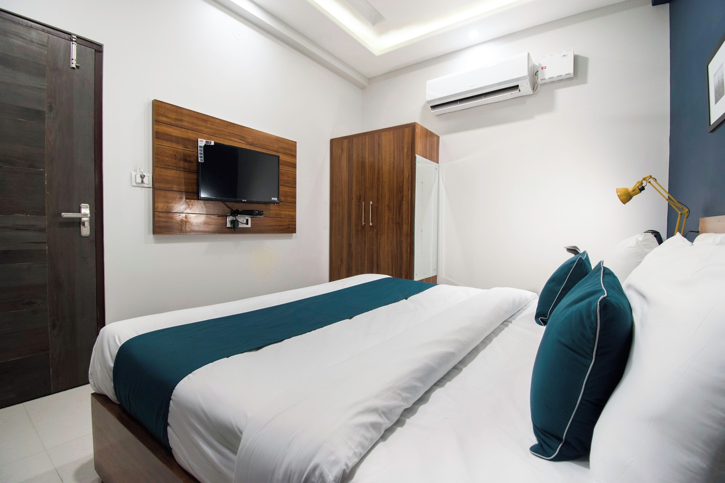 A hotel room with a bed, a television mounted on a wooden panel, an air conditioning unit, and a desk lamp.