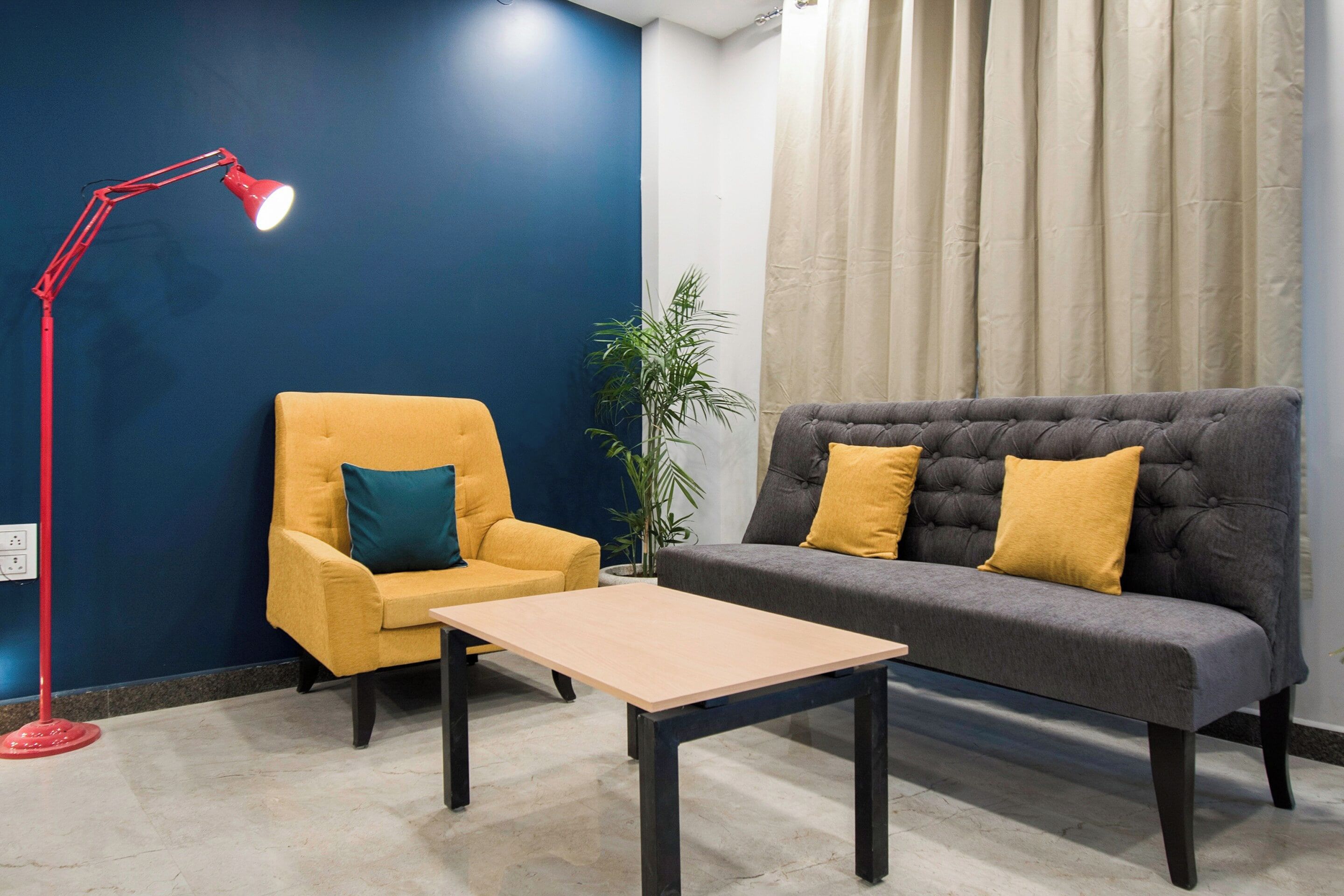 A room with a blue wall, a yellow armchair, a grey sofa with yellow cushions, a red floor lamp, and a potted plant.