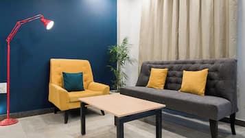 A room with a blue wall, a yellow armchair, a grey sofa with yellow cushions, a red floor lamp, and a potted plant.