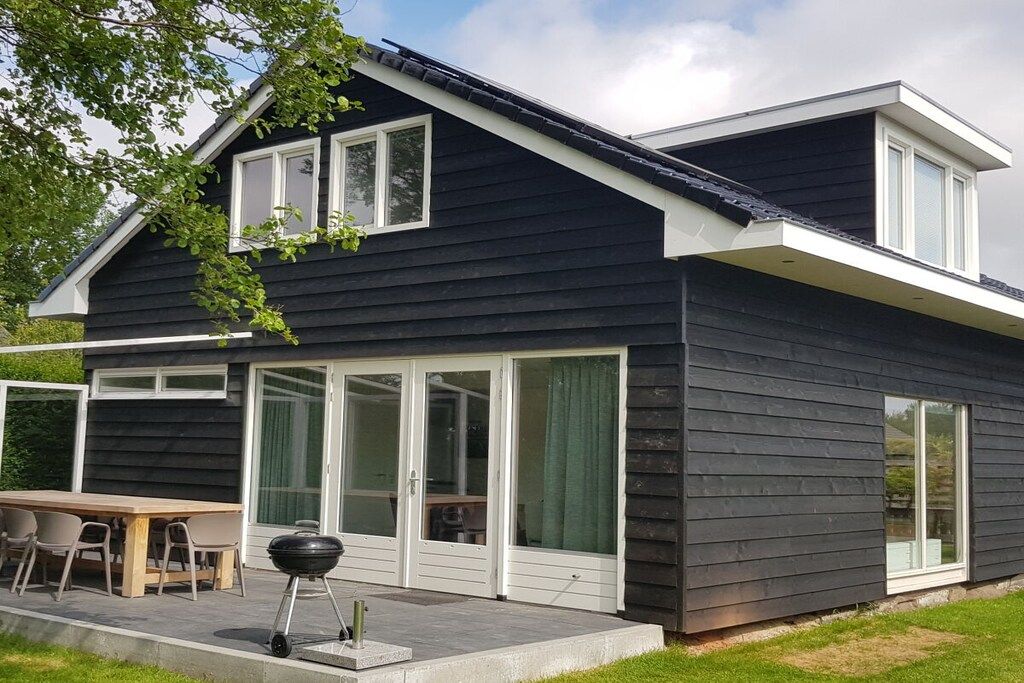 A modern two-story house with a dark exterior, large sliding glass doors, and a patio with outdoor furniture.