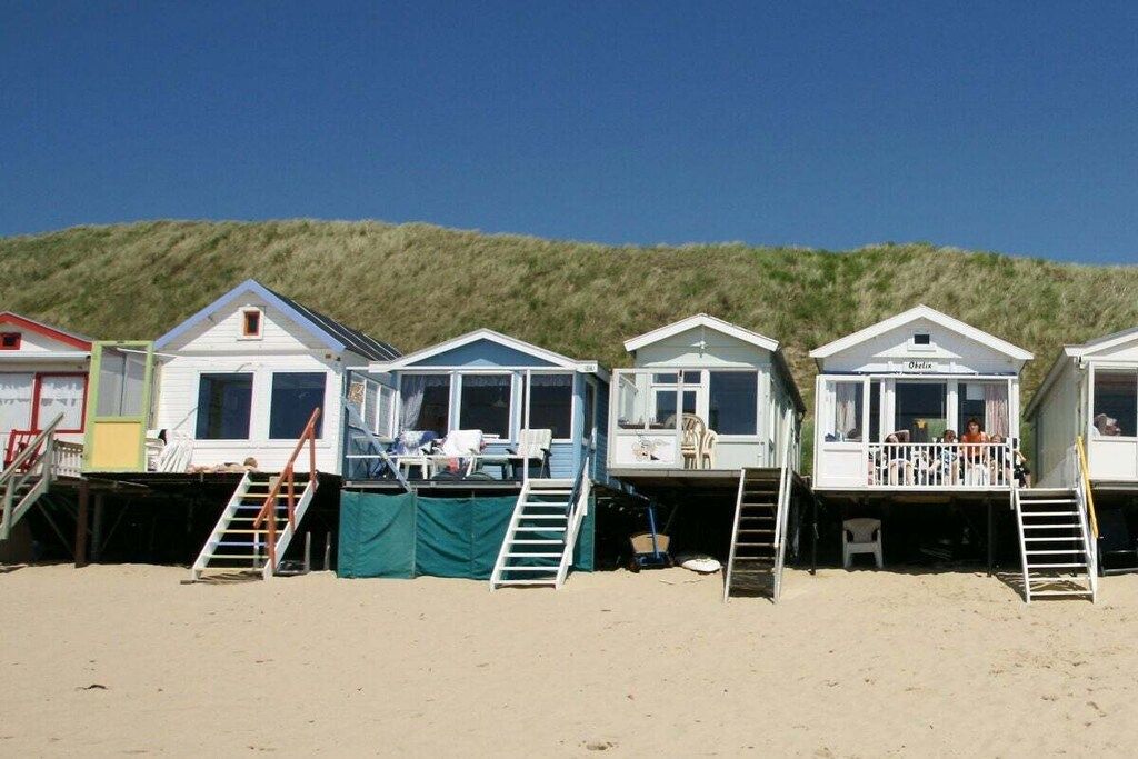 Una hilera de casas de playa con escaleras exteriores que dan al acceso, ubicadas en una playa arenosa con una colina cubierta de hierba al fondo.
